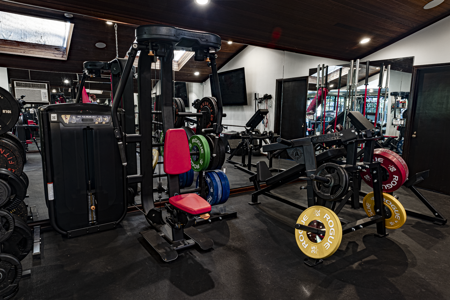 Luxury home gym with Matrix and Arsenal Strength machines, color-coded Rogue bumper plates, and a mirrored wall under a wood-paneled ceiling with skylights